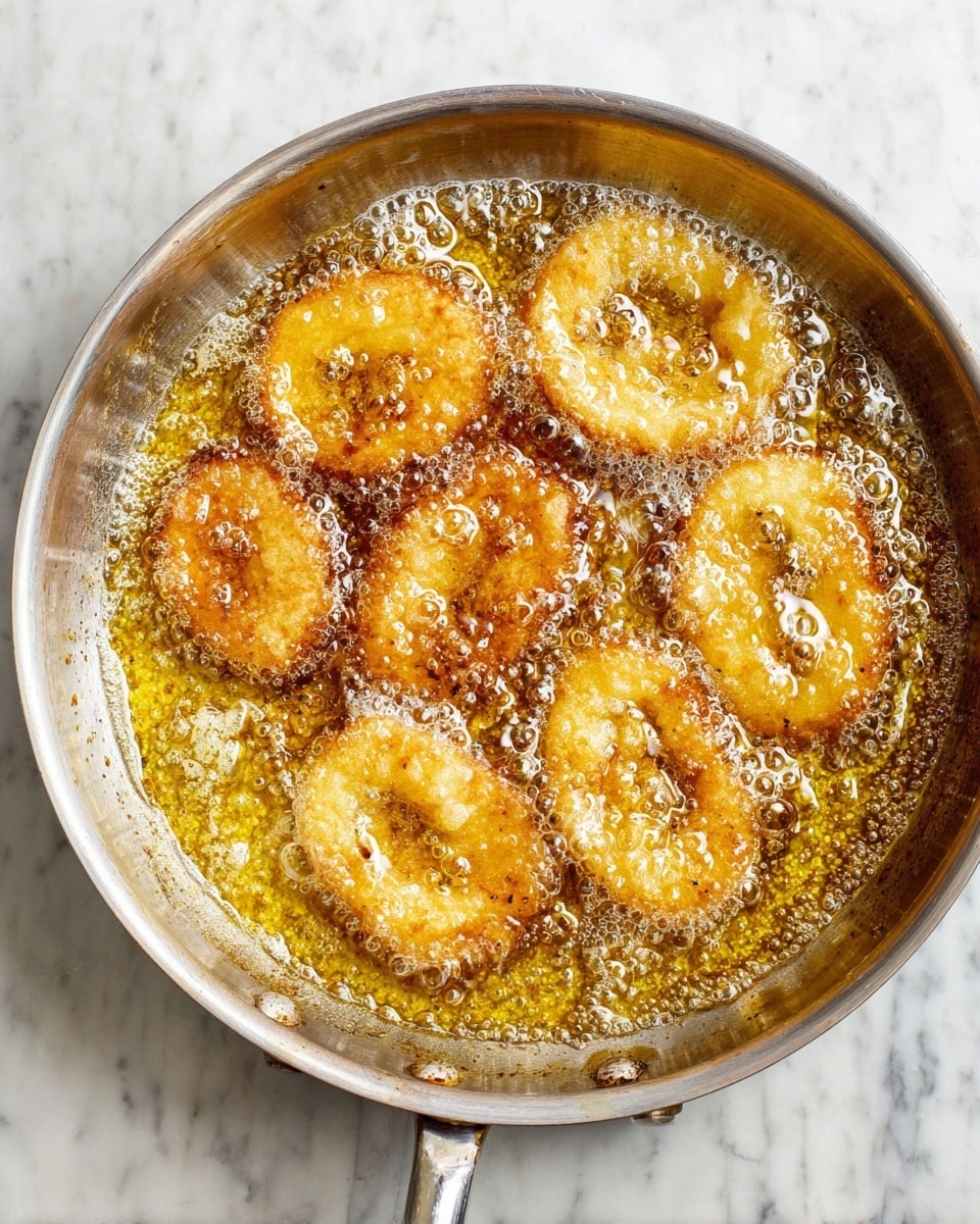 A metal pan filled with bubbling golden oil shows several round and oval light brown rings frying inside, the rings have a slightly crispy texture with some darker brown spots. The oil is clear with many small bubbles swirling around the rings, creating a hot frying effect. The pan is sitting on a white marbled surface, and the handle is visible on the right side. The photo taken with an iphone --ar 4:5 --v 7