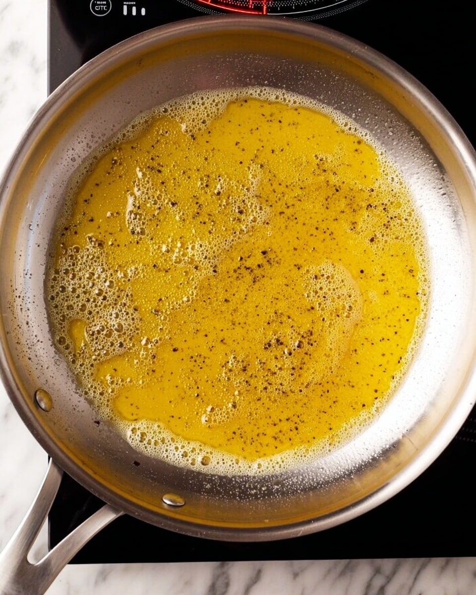 A large silver pan filled with cooked spaghetti noodles, lightly coated in a glossy sauce with visible black pepper flakes scattered throughout. The spaghetti strands are tangled and piled evenly inside the pan, which sits on a white marbled surface. The pan has two shiny metal handles on either side, and the stove display in the background shows a cooking time. photo taken with an iphone --ar 4:5 --v 7