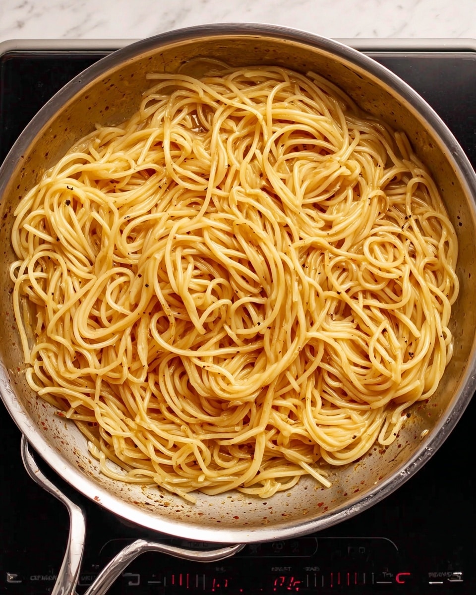 A close-up view of a shiny silver frying pan filled with melted yellow butter mixed with small black pepper specks bubbling gently across the surface, held on a black stove top with digital controls visible at the top, all against a white marbled countertop. The pan has two handles, one on each side, and reflects the light softly on its smooth surface photo taken with an iphone --ar 4:5 --v 7
