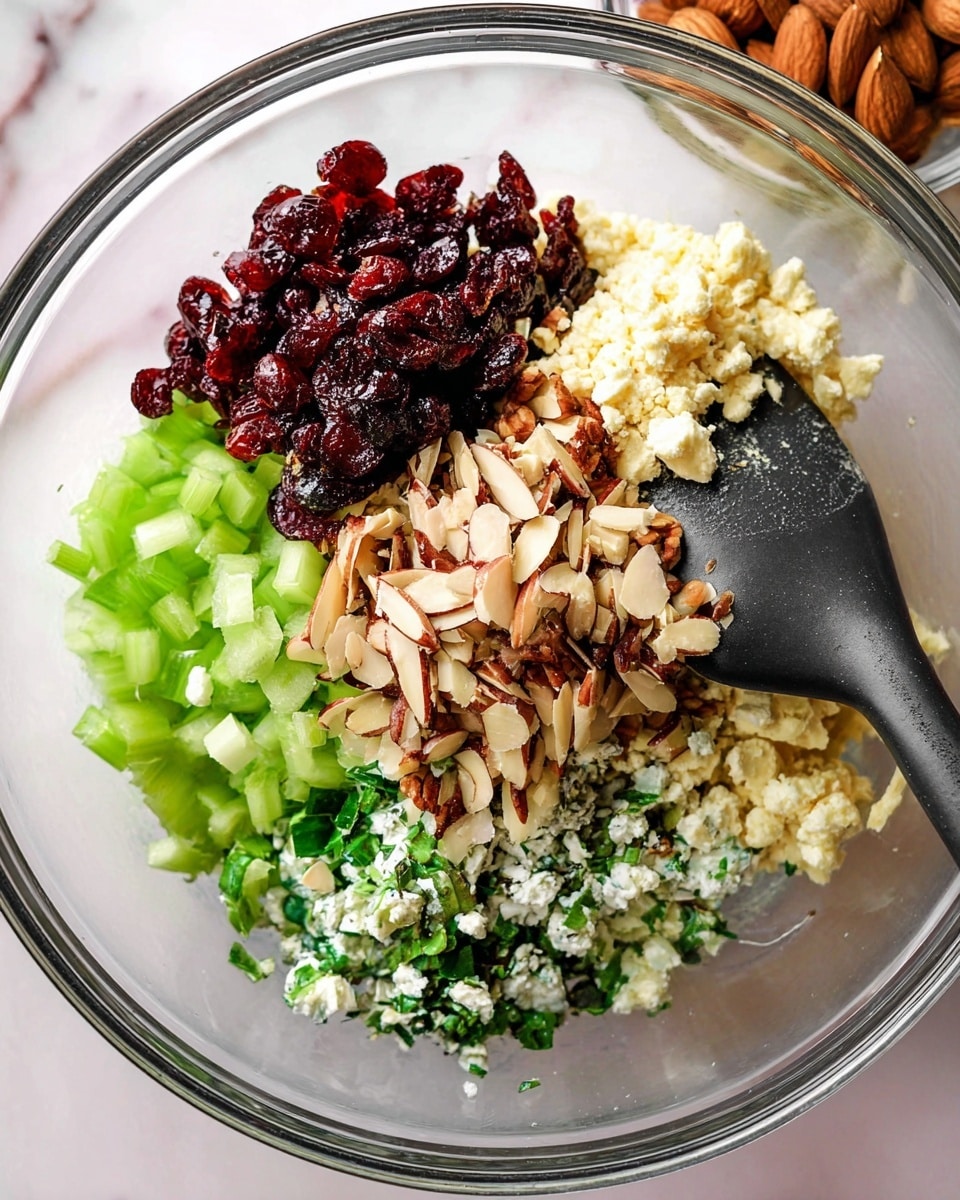 The image shows a clear glass bowl with six visible layers of ingredients arranged separately but close together: dark red dried cranberries on the left, light green sliced celery below them, dark green chopped herbs beneath the celery, golden cooked onions on the right, creamy white crumbled cheese next to the onions, and toasted sliced almonds on top in the center. A black spatula is partially inside the bowl, touching the cooked onions. The bowl is placed on a white marbled surface. Photo taken with an iphone --ar 4:5 --v 7