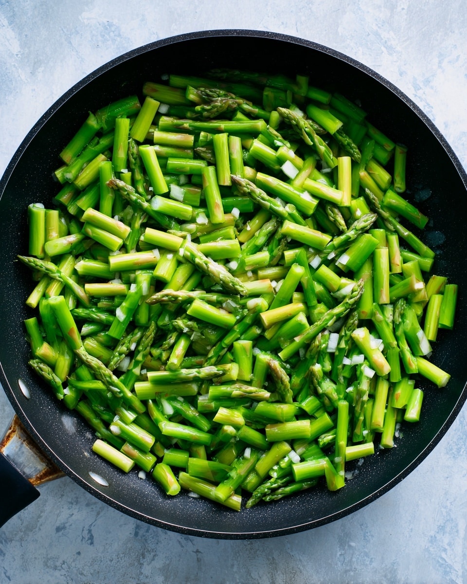 In a large deep black pan, small bright green asparagus pieces are spread evenly across the surface. The asparagus is cut into thick, short cylindrical shapes, showing a fresh green color with hints of pale light green at the ends. Bits of white chopped garlic are also visible scattered among the green asparagus. The pan sits on a cooking surface with a white marbled texture beneath it. photo taken with an iphone --ar 4:5 --v 7