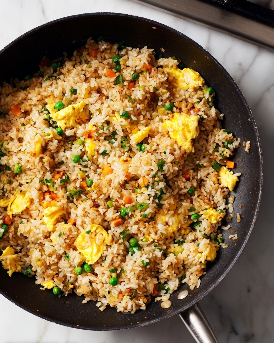 A close-up view of a black frying pan filled with cooked fried rice mixed with scrambled eggs. The rice grains are light brown with some darker spots, showing a cooked texture, while soft yellow scrambled egg pieces are spread evenly through the dish. The frying pan sits on a white marbled surface, with the metallic stove burner partially visible behind it. The lighting softly highlights the texture of the rice and eggs. Photo taken with an iphone --ar 4:5 --v 7