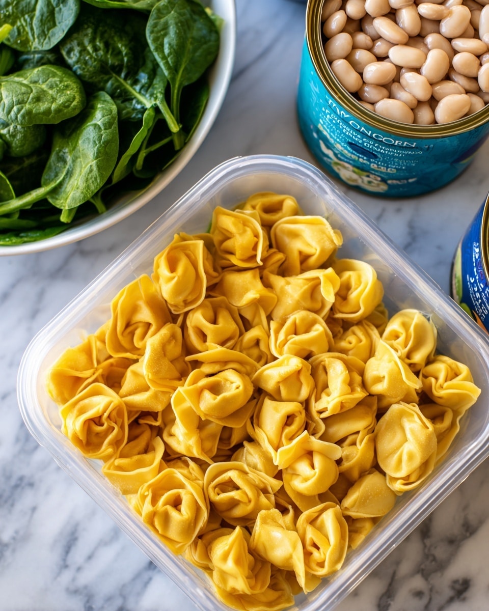 The image shows a clear plastic container filled with many pieces of yellow tortellini pasta, each with a slightly wrinkled texture and folded into small rings, laying closely together. Behind the container, there is a white bowl with dark green spinach leaves that look fresh and vibrant. To the right, there is a can with a blue label featuring white beans and some text. The background is a white marbled texture. photo taken with an iphone --ar 4:5 --v 7