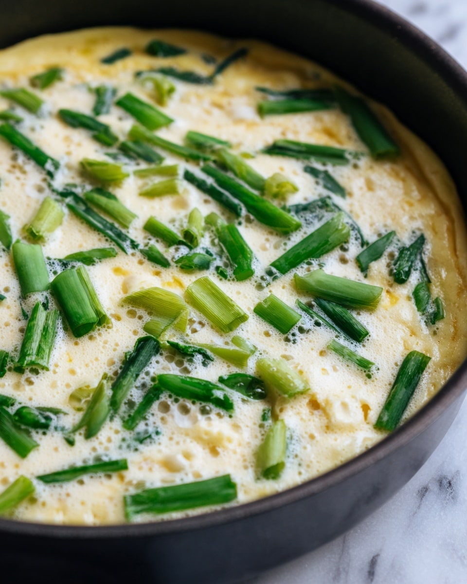 A white plate holds a round, golden-brown pancake with green pieces visible inside, cut into several square pieces. One square piece is held by silver chopsticks above a small clear glass bowl filled with dark soy sauce mixed with chopped green onions, creating a contrast of colors. The pancake's surface is slightly crispy with charred marks, and the white marbled texture below adds brightness to the scene. photo taken with an iphone --ar 4:5 --v 7