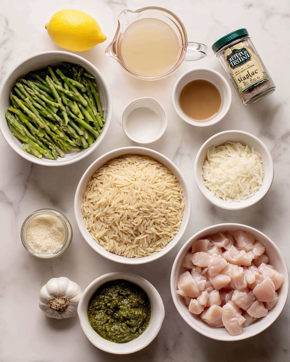 The image shows a top view of several white bowls and a clear measuring cup arranged on a white marbled surface. The top right bowl contains light pink cubed raw chicken pieces. Below it, a large bowl is filled with dry, pale beige orzo pasta. To the left of the orzo, another medium bowl holds chopped green asparagus with dark green tips. Above this bowl, there is a small bowl filled with thick, dark green pesto sauce. Next to the pesto, a clear measuring cup contains a light brown broth. Above the broth is a small white bowl with a white liquid, likely cream or milk. To the left of the cream bowl is a yellow lemon and two garlic cloves. Above the chicken bowl, there is a glass jar labeled