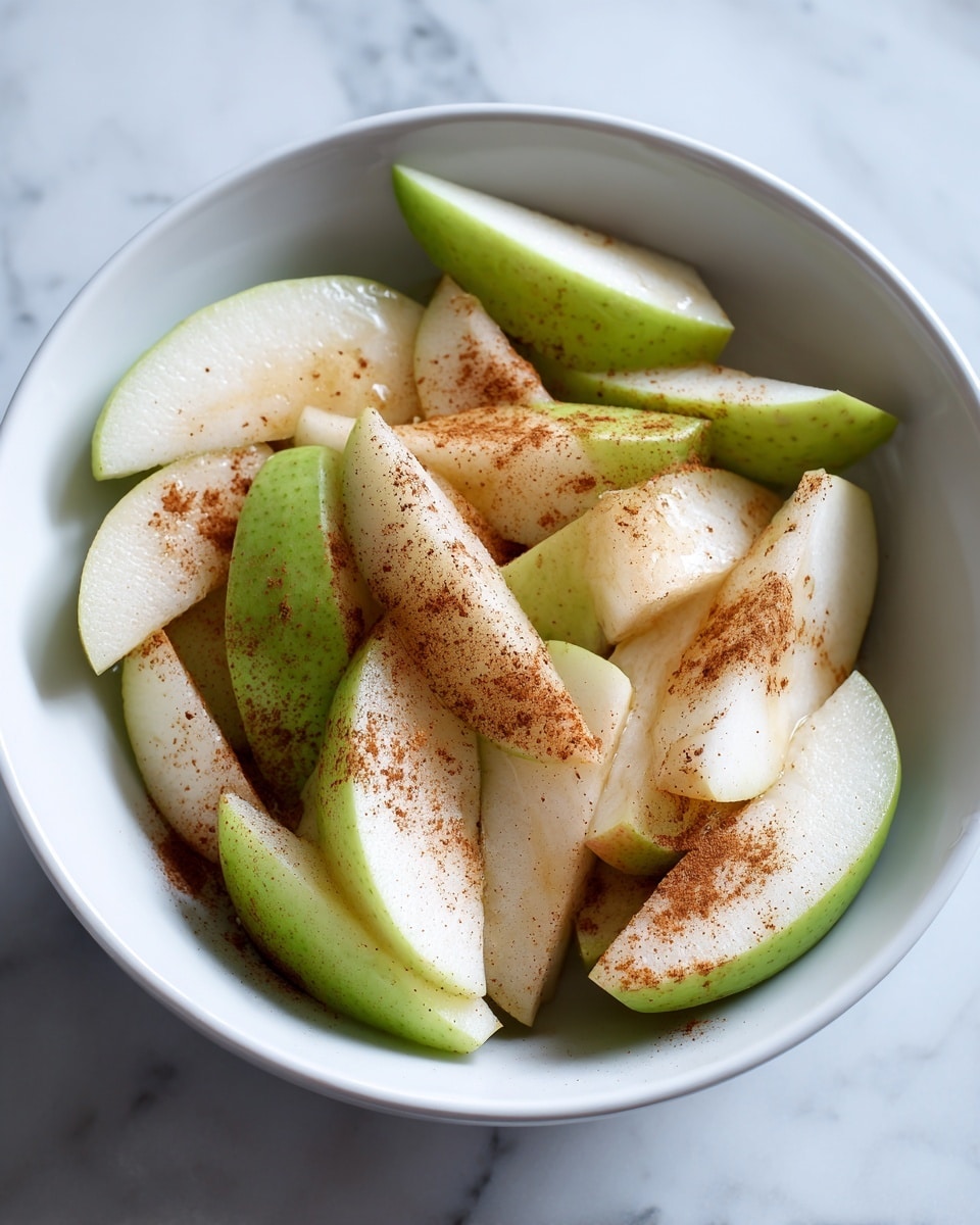 The image shows a white bowl filled with several slices of apples and pears. The apples are mostly green with a hint of red skin, and their smooth flesh looks juicy and fresh. The pear slices are light green to white, slightly translucent with a firm texture. Both the apple and pear slices are sprinkled unevenly with brown cinnamon powder and a lighter beige powder, adding a warm contrast to the pale fruit colors. The fruits appear moist, with a few drops of liquid visible, and the bowl sits on a surface with a white marbled texture. Photo taken with an iphone --ar 4:5 --v 7