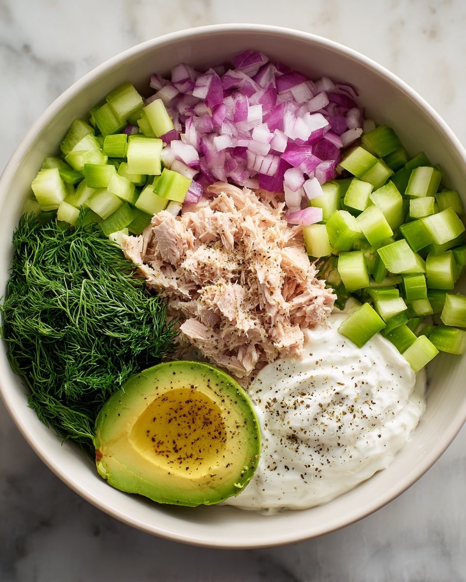 A white bowl contains six separate layers arranged in a circle. At the top right, there are small green celery cubes. Next to the celery, moving clockwise, finely chopped red onions are placed. Below the onions, a thick bunch of fresh green dill fills the space. At the bottom left, there is half of an avocado with a smooth green texture and some black pepper on top. Above the avocado, a creamy white layer of yogurt or sour cream is placed. In the center, light brown shredded tuna is situated, sprinkled lightly with black pepper and seasoning. The bowl sits on a white marbled surface. Photo taken with an iphone --ar 4:5 --v 7
