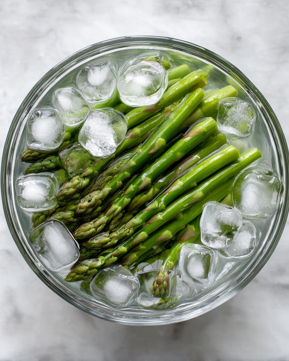 A clear glass bowl filled with bright green asparagus spears lying flat inside. The asparagus is covered with clear water and many irregularly shaped ice cubes floating on top, creating a fresh and cold look. The bowl is placed on a white marbled surface that adds a clean and simple background to the scene. photo taken with an iphone --ar 4:5 --v 7