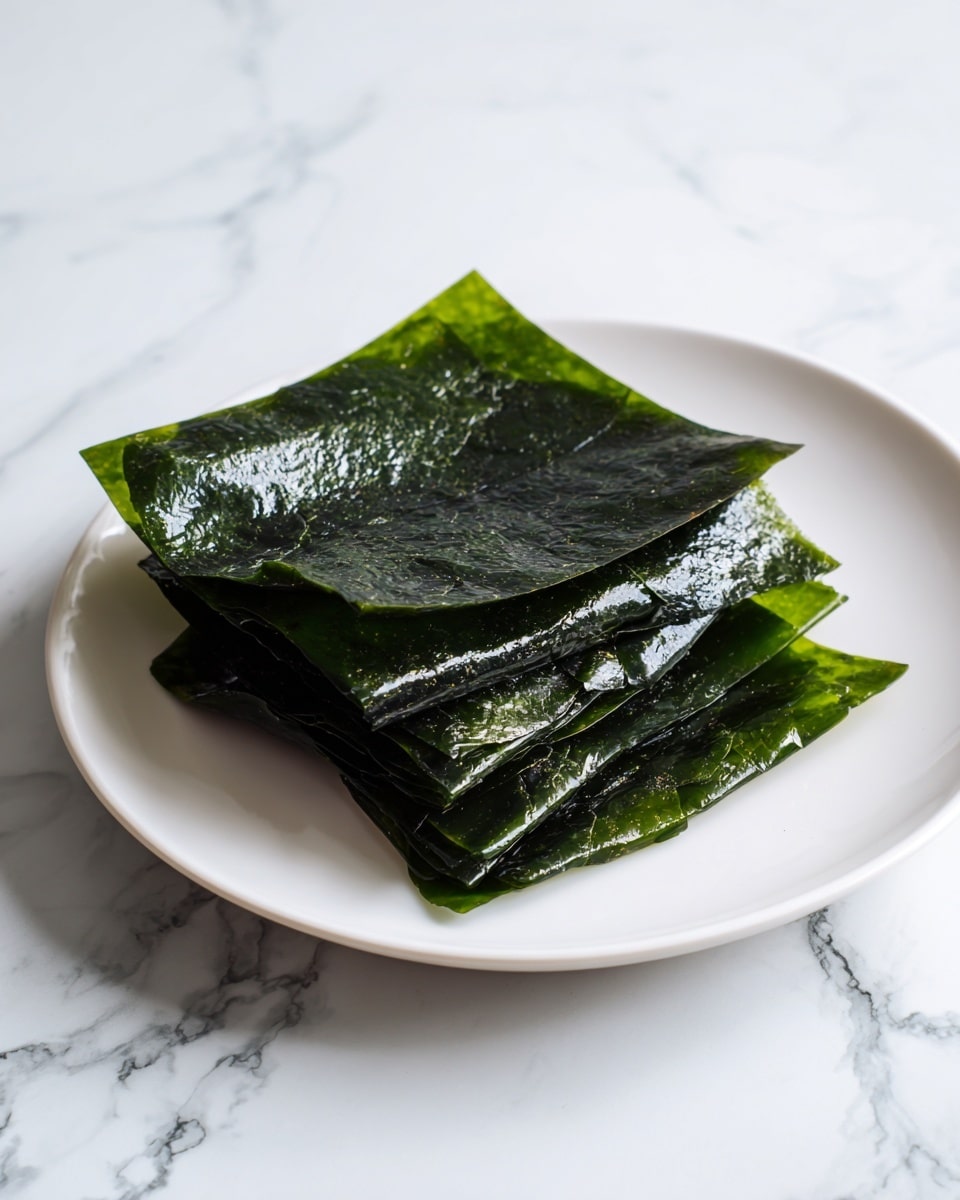 A small stack of dark green, shiny seaweed sheets is neatly placed on a white plate, showing about four thin, textured layers with a slightly crinkled surface. The seaweed is glossy and the white marbled table surface underneath adds a clean look to the image. photo taken with an iphone --ar 4:5 --v 7