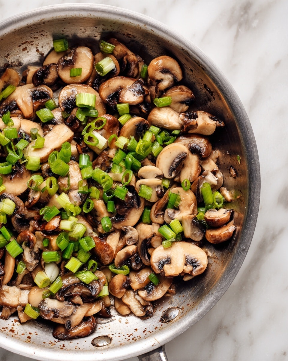 The image shows a close-up of a stainless steel pan filled with cooked mushrooms and chopped green onions. The mushrooms are sliced and whole in light brown and tan colors, with some darker brown gills visible. The green onions are bright green and scattered on top and between the mushrooms. The pan’s inside surface is slightly shiny with a few brown spots from cooking, all on a white marbled texture background. photo taken with an iphone --ar 4:5 --v 7