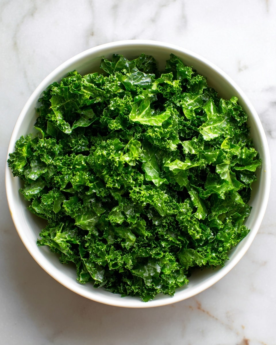 A white bowl filled with a single layer of finely chopped bright green kale leaves that look fresh and slightly wet, sitting on a white marbled surface. The kale pieces are curly and cover the entire inside of the bowl evenly, with some bits close to the edge. Photo taken with an iphone --ar 4:5 --v 7