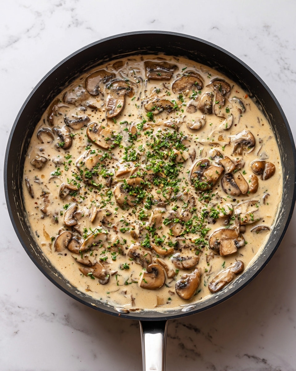 A black pan filled with creamy mushroom sauce, with many light beige sauce layers mixed with sliced light brown mushrooms scattered throughout. The top has a small sprinkle of green chopped herbs in the middle for decoration. The pan sits on a white marbled surface. photo taken with an iphone --ar 4:5 --v 7