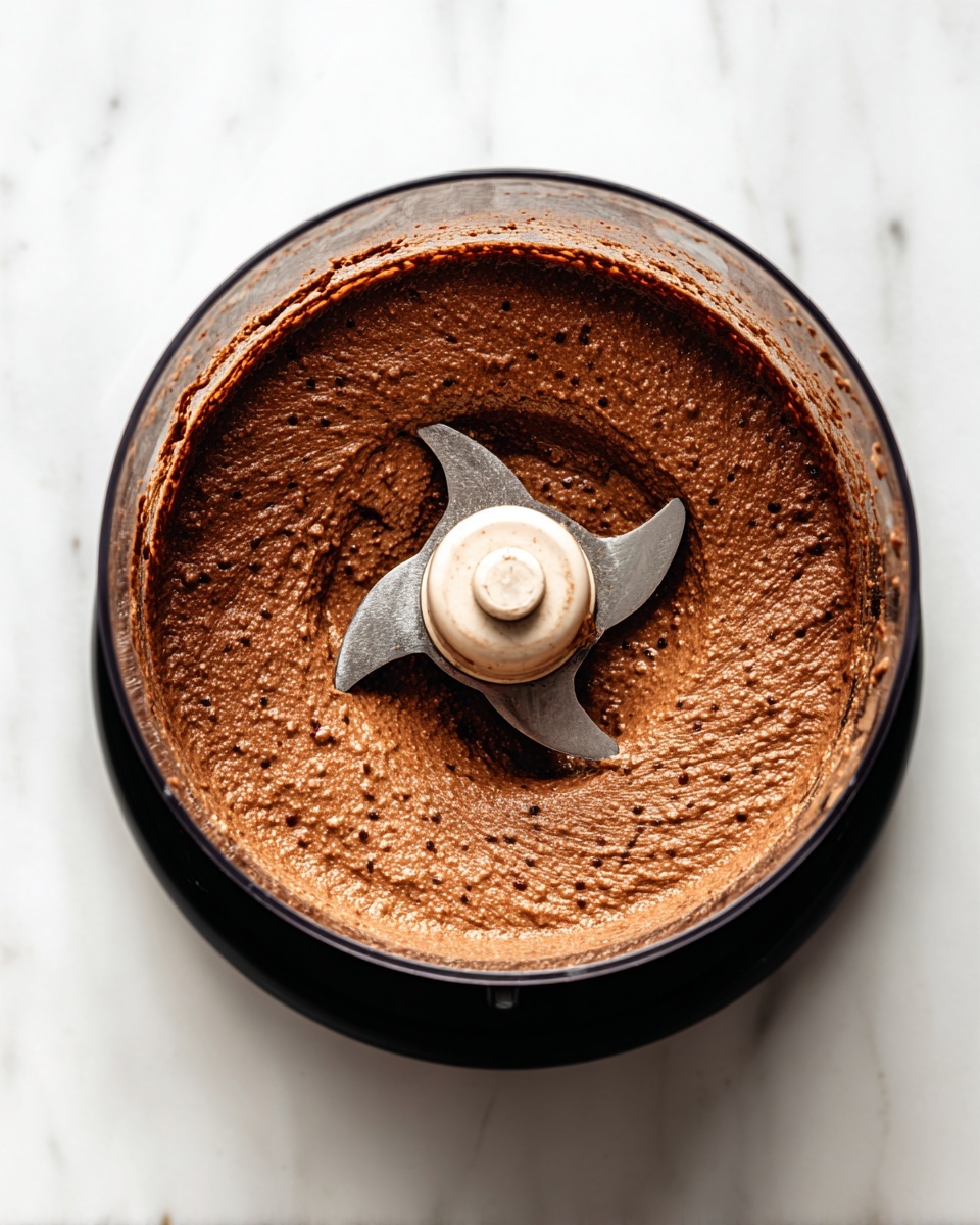 A top view of a clear food processor bowl containing a thick brown paste with tiny specks, spread evenly along the sides and bottom around the metal blades in the center. The bowl is on a black base, placed on a white marbled surface. The texture of the paste looks smooth but dense, covering the blades partially with some residue on the sides. Photo taken with an iphone --ar 4:5 --v 7