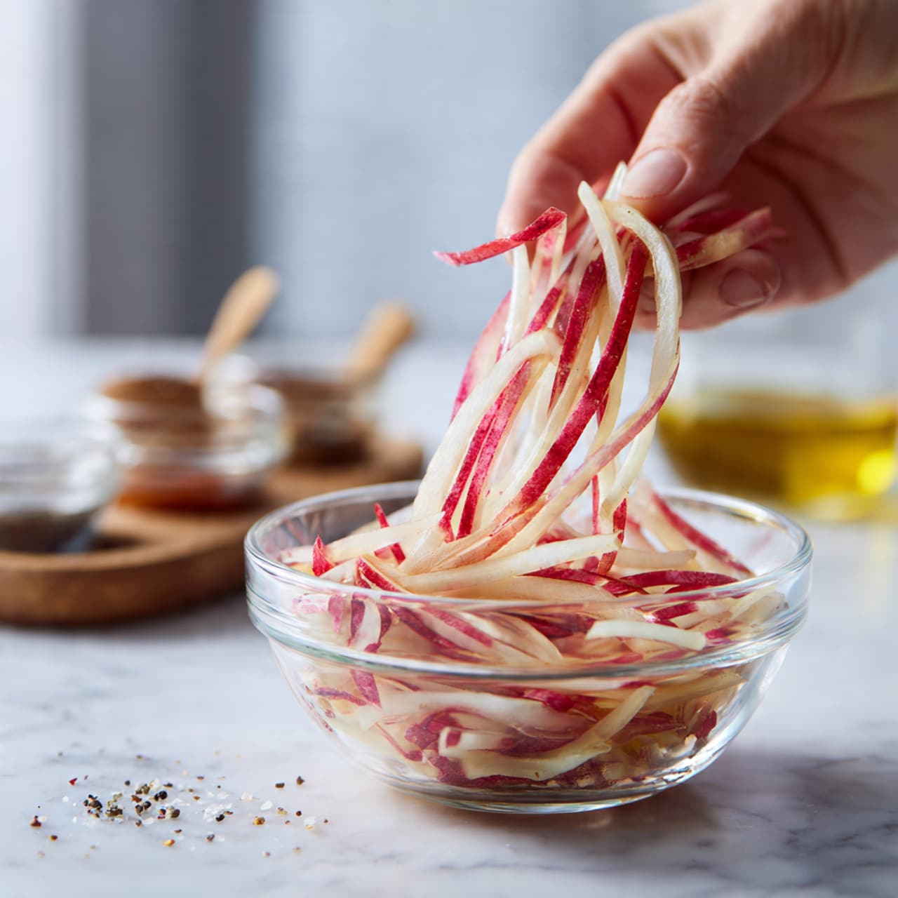 In a clear glass bowl on a white marbled surface, there is one main layer of light pink salmon flakes with charred black pepper spots spread evenly around the inside. In the middle, a smooth dark red sauce pool is topped by a swirl of creamy pale yellow sauce being poured in from above, creating a soft texture contrast. The bowl shows no other layers visible beneath the salmon. Photo taken with an iphone --ar 4:5 --v 7