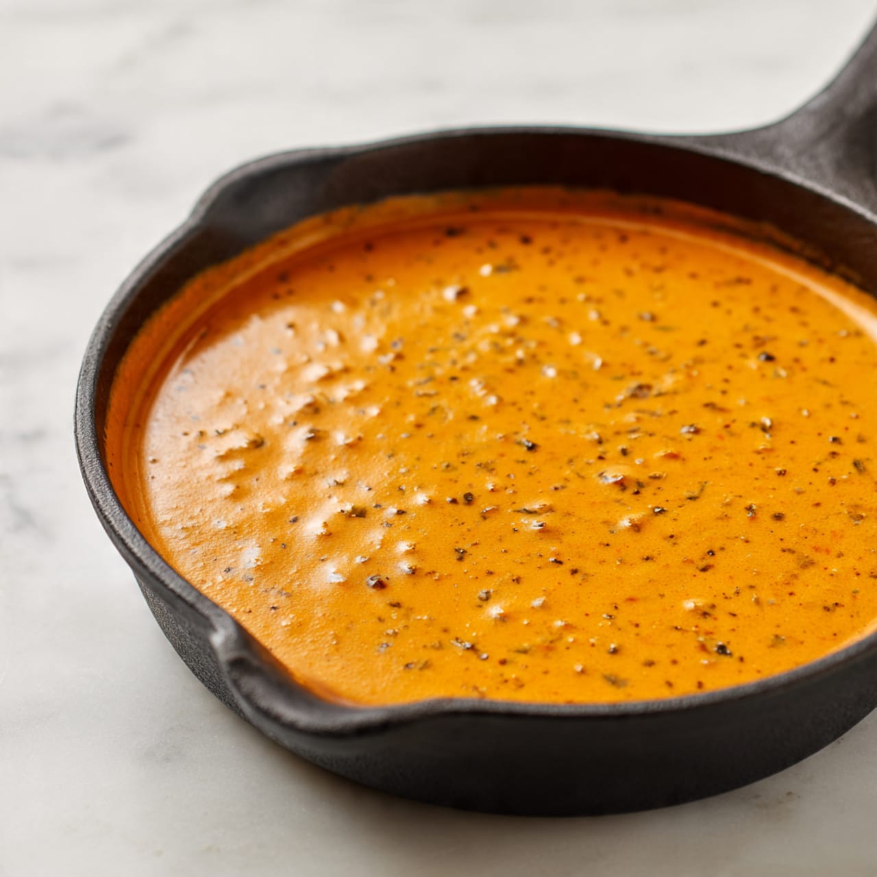 A white bowl holds two main layers: the bottom layer is plain white rice with a soft and fluffy texture, and the top layer is creamy orange sauce with pieces of chicken. The chicken pieces are light brown and covered well by the sauce, with small green herb pieces scattered on top. A silver spoon rests on the rice inside the bowl. The bowl is set on a white marbled surface with green herb leaves around it. Photo taken with an iphone --ar 4:5 --v 7