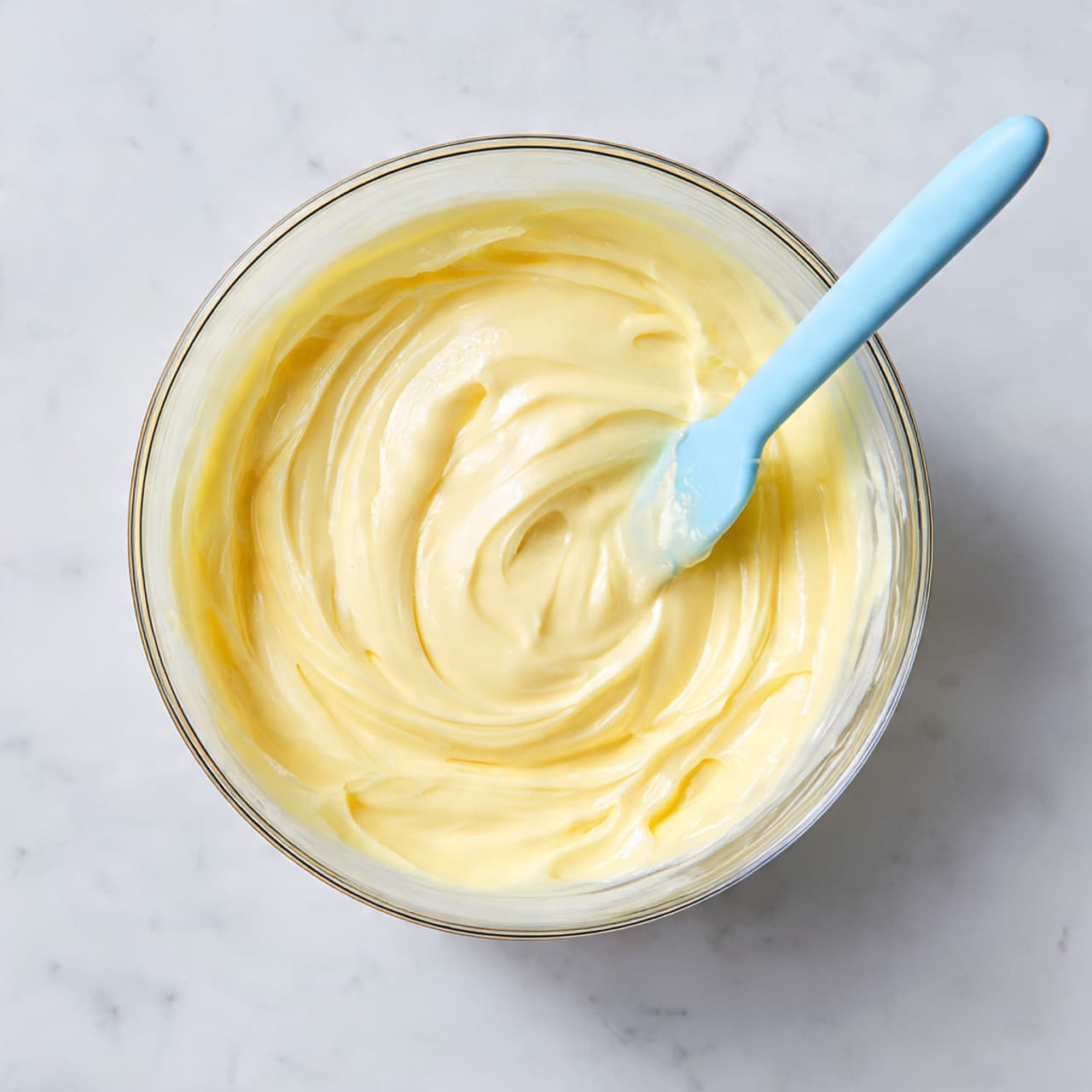 A clear glass bowl holds a smooth, pale yellow mixture with a creamy texture. Inside the bowl, a light blue silicone spatula is partially dipped into the mixture, creating soft swirls. The bowl is placed on a white marbled surface, giving a clean and simple background. The overall scene is softly lit, highlighting the glossy, gentle shine of the creamy mixture. photo taken with an iphone --ar 4:5 --v 7