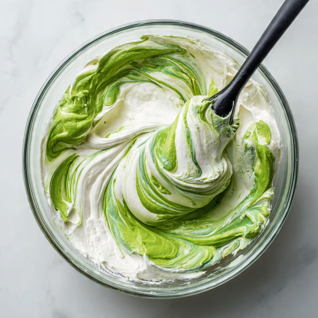 A close-up view of a glass bowl filled with a swirling green and white creamy mixture. The green part is thick and smooth, mixed with white cream creating a marbled pattern. A black spatula lifts some of the mixture, showing its soft and fluffy texture as it folds back into the bowl. The mixture looks airy and light, with shine and smoothness visible in the folds. The bowl sits on a white marbled surface. photo taken with an iphone --ar 4:5 --v 7