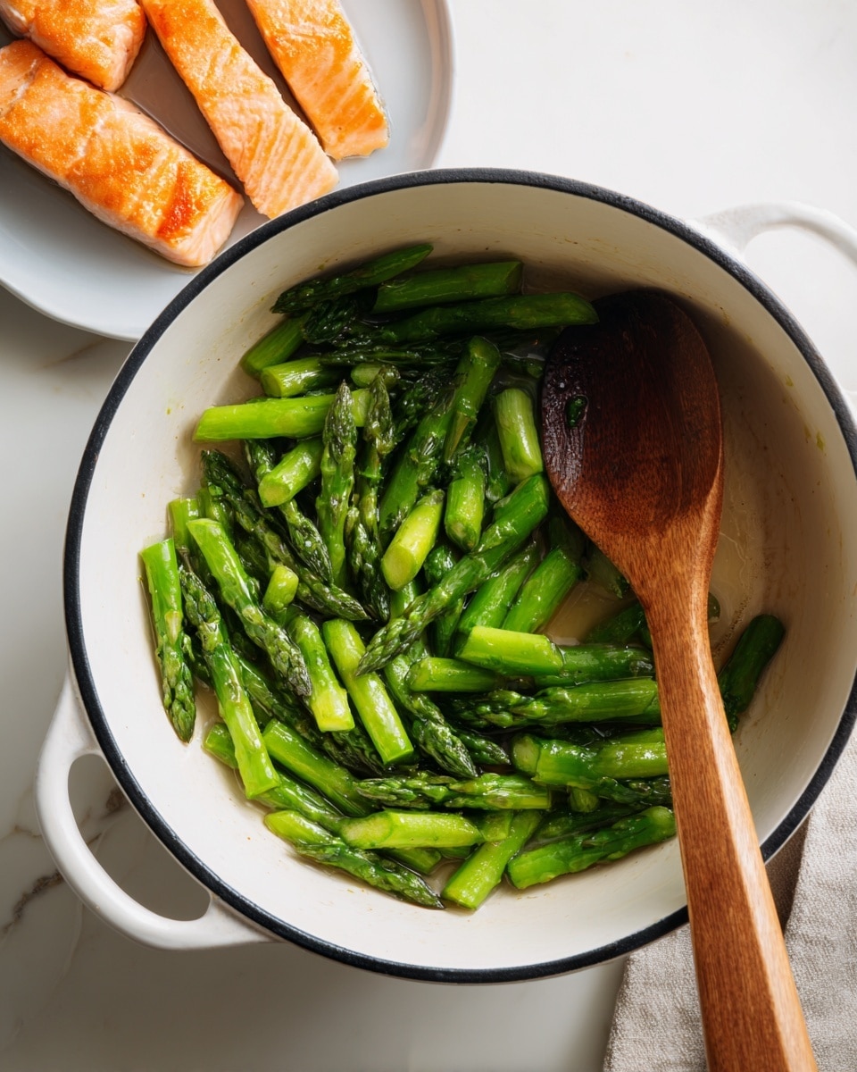 The image shows a white pot with a black rim filled with bright green asparagus pieces, lightly sautéed and slightly shiny with oil. The asparagus sticks are cut into medium-length pieces, spread somewhat loosely inside the pot. A light wooden spoon rests inside the pot with its handle extending outwards on a white marbled surface. Above the pot, there is a white plate holding several pieces of golden-brown cooked salmon arranged in a casual manner. The overall scene is bright with a clean, white marbled background and soft natural lighting. photo taken with an iphone --ar 4:5 --v 7