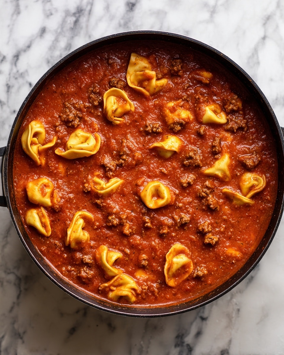 The image shows a pot filled with a rich red sauce mixed with meat and tortellini pasta. The sauce is thick and smooth, covering the ingredients evenly. The tortellini are yellowish with some parts darker from cooking, scattered throughout the sauce. Small pieces of brown ground meat are visible inside the sauce, creating texture and color contrast. The pot edges are black, creating a dark frame around the warm food. The background is a white marbled texture. Photo taken with an iphone --ar 4:5 --v 7