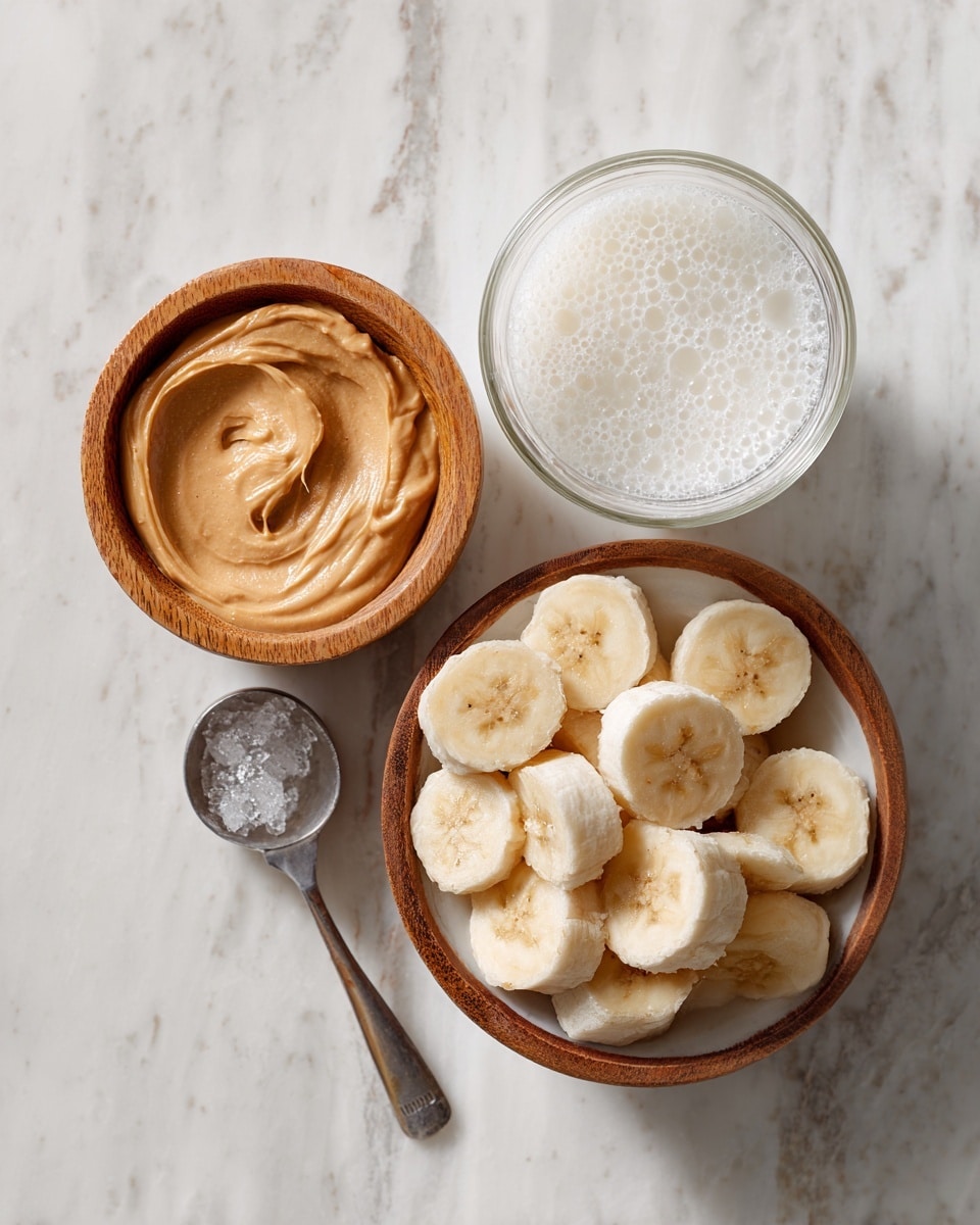 The image shows three small bowls on a white marbled surface: the first bowl is wooden and filled with a smooth, light brown peanut butter, the second bowl is white and contains frozen banana slices covered lightly in ice crystals, and the third is a clear glass bowl with white milk slightly frothy on top. A silver measuring spoon is placed near the bowls. photo taken with an iphone --ar 4:5 --v 7