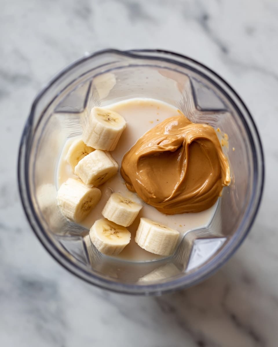 The image shows the inside of a blender from above, with three main layers visible. At the bottom, there is a small amount of light brown liquid, which looks like milk or a similar drink. On top of this, there are several small, light cream-colored banana pieces scattered around. Finally, a large dollop of smooth, light brown peanut butter sits on one side, slightly overlapping the banana pieces. The blender container is clear, and the background is a white marbled surface. photo taken with an iphone --ar 4:5 --v 7