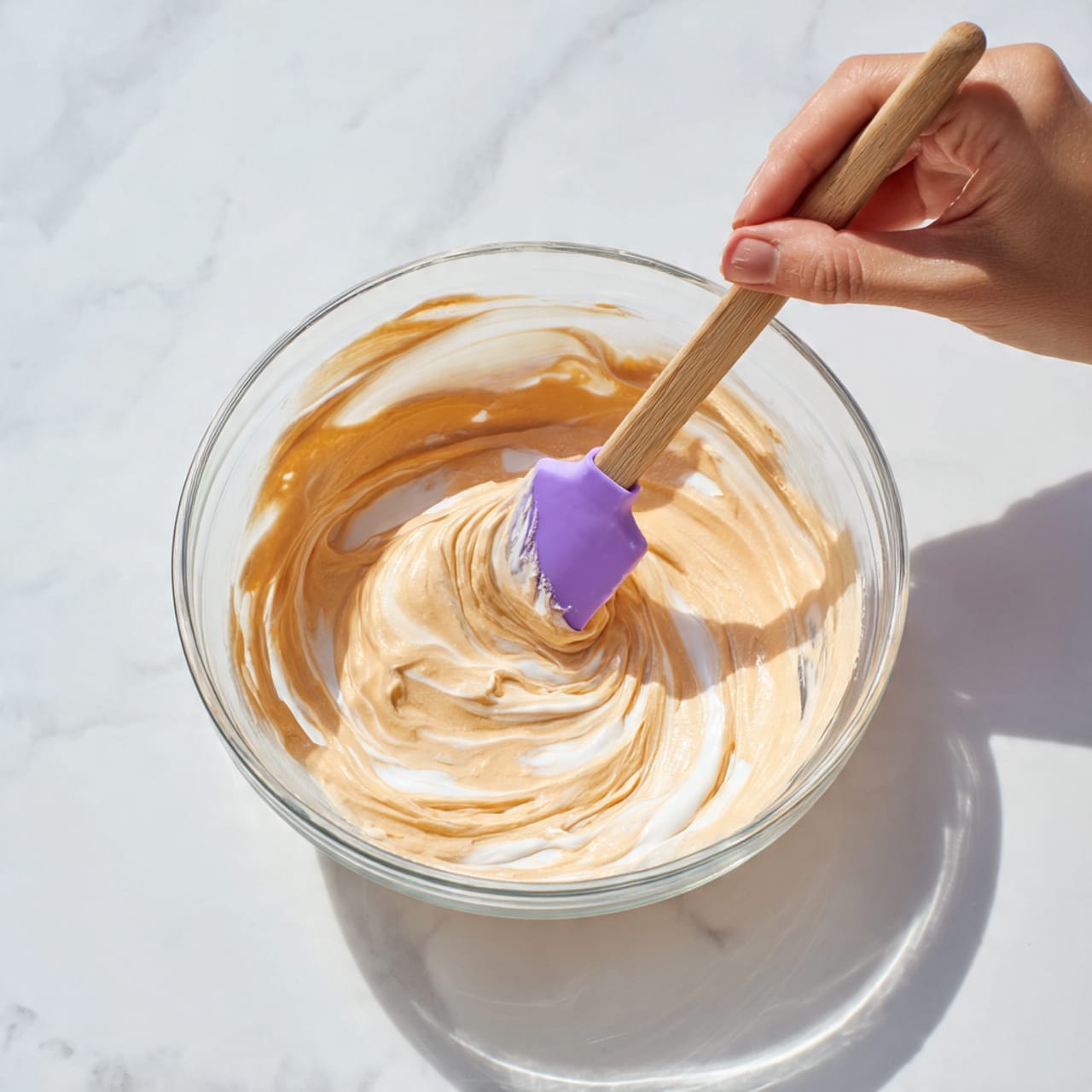 A clear glass bowl sits on a white marbled surface filled with a creamy mixture that has light orange and tan swirls throughout, showing the smooth, thick texture being stirred. A purple spatula with a wooden handle is held by a woman's hand, lifting a dollop of the swirled creamy mix above the bowl, creating soft peaks and curved lines in the mixture inside the bowl. The lighting is bright and natural, highlighting the shiny and soft texture of the mix. photo taken with an iphone --ar 4:5 --v 7