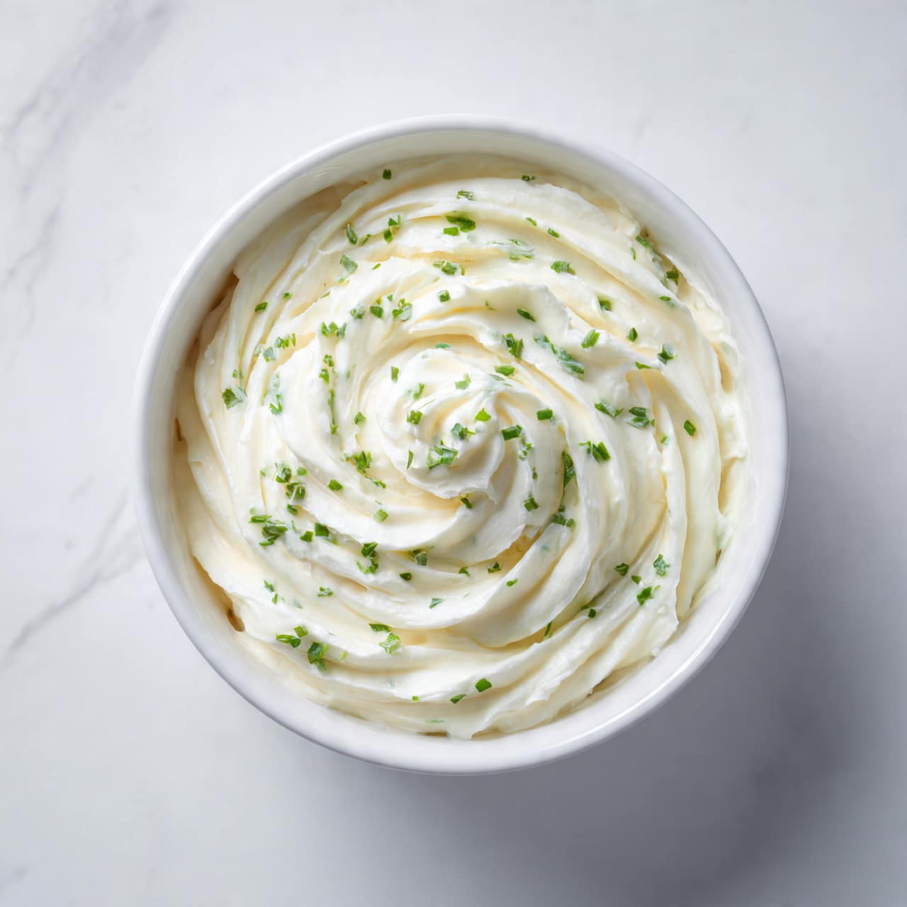 A white bowl sits on a white marbled surface, filled with one smooth layer of creamy white spread mixed with small green herb pieces scattered evenly throughout. The spread has soft, rounded swirls creating light texture on the surface. The bowl is centered, showing a close view of the creamy, slightly whipped appearance of the spread. Photo taken with an iphone --ar 4:5 --v 7