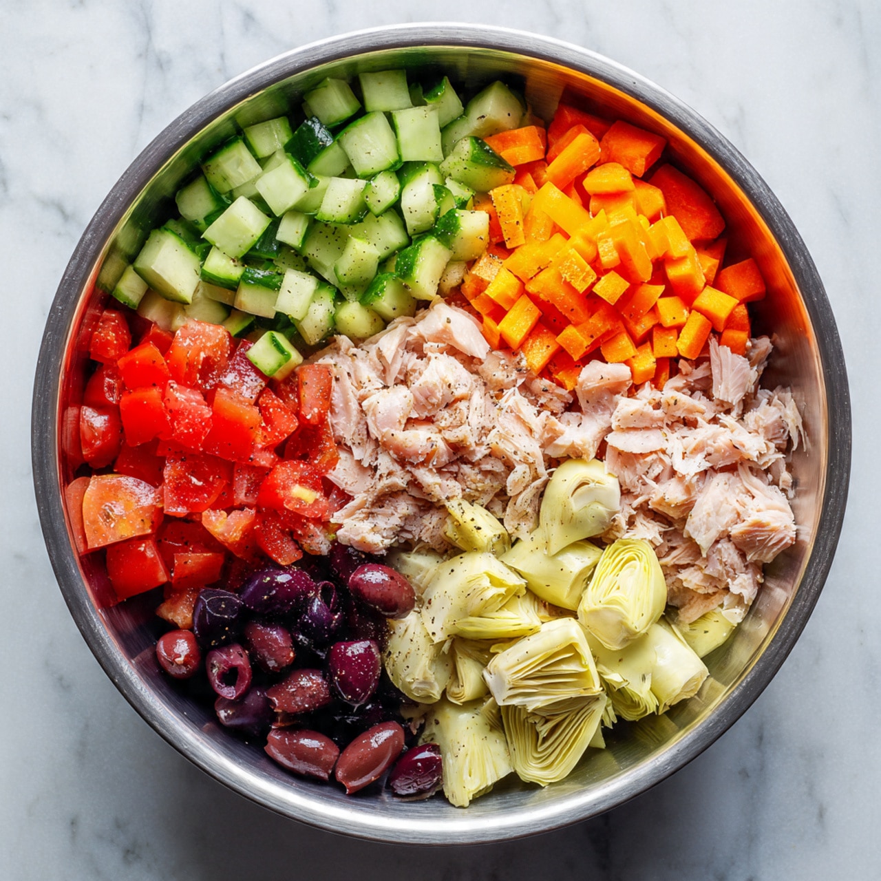 A large silver metal bowl contains seven visible layers of diced and chopped ingredients, each arranged in separate sections inside the bowl. Starting from the top left and moving clockwise, there are light green pieces of cucumber, small bright orange diced carrots, halved bright red cherry tomatoes, shredded pale beige tuna, dark purple sliced olives, bright red diced bell peppers, and light beige artichoke hearts. Some black pepper is sprinkled over several ingredients, adding small black specks. The bowl rests on a white marbled surface. photo taken with an iphone --ar 4:5 --v 7