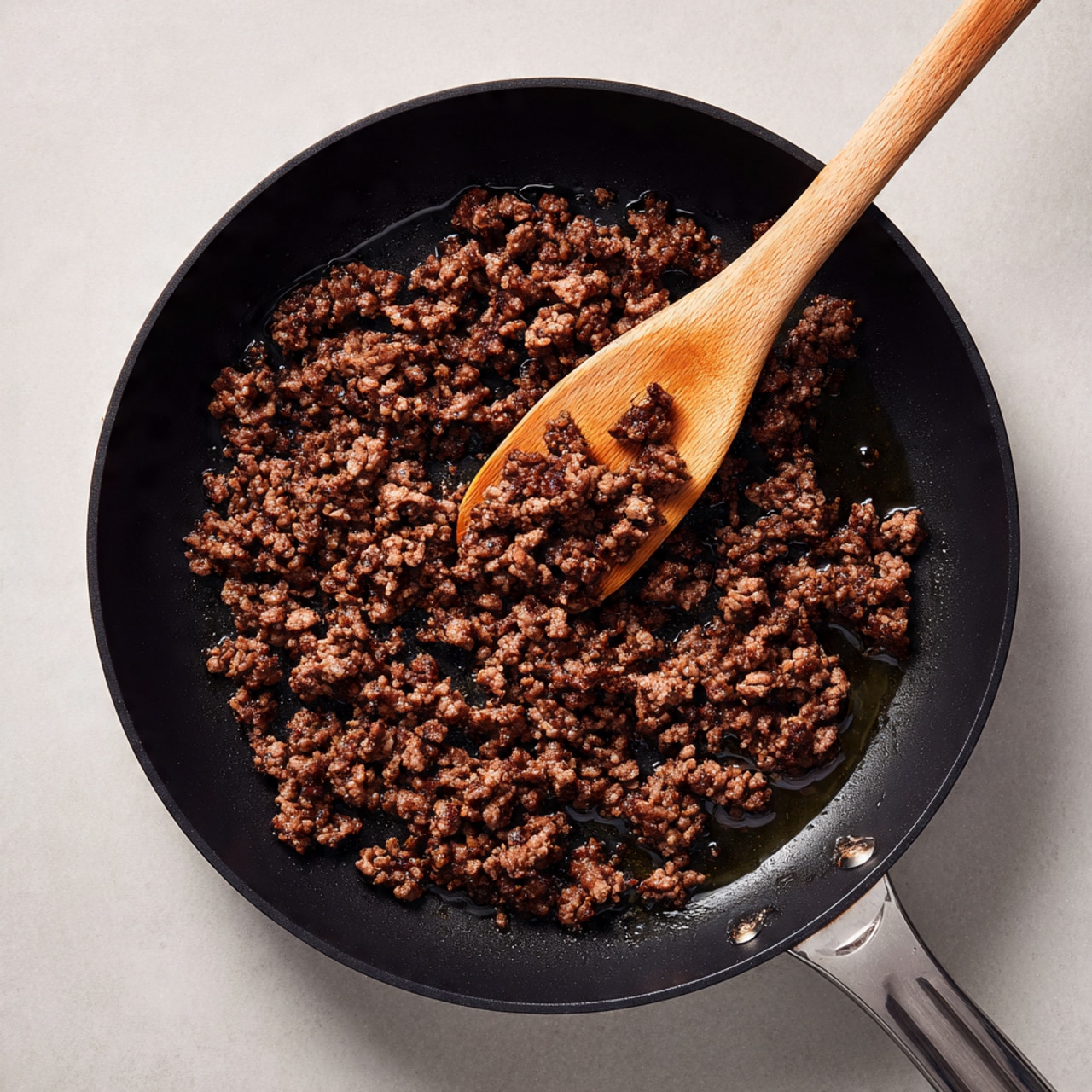 A close-up view of cooked ground beef in a black frying pan. The meat is broken into small pieces, browned with some parts showing a slightly pinkish tint, and surrounded by a thin layer of oil glistening under the light. A wooden spatula is partially visible, resting on the edge of the pan and slightly stirring the beef. The pan surface is dark with a few drops of oil scattered around. The background surface is not visible, focusing fully on the pan and cooked beef. photo taken with an iphone --ar 4:5 --v 7