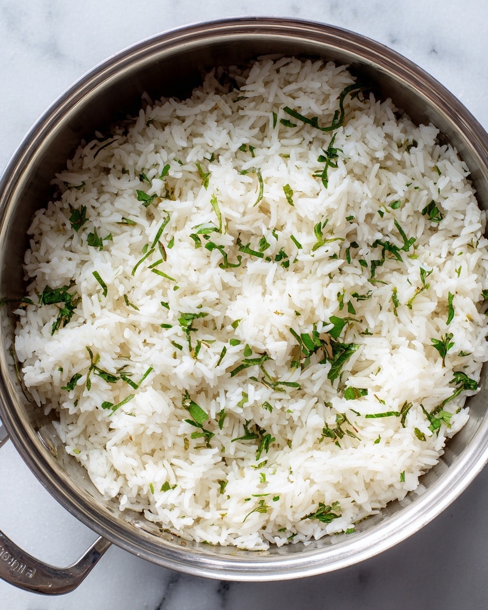 A close-up view of cooked white rice mixed with small green herb pieces spread evenly throughout, inside a shiny metal pot placed on a white marbled surface. The rice grains look soft and fluffy with a slightly wet texture, and the bright green herbs add a fresh contrast to the white rice. The pot’s edge is visible above the rice, showing its metal rim and handle at the bottom. photo taken with an iphone --ar 4:5 --v 7
