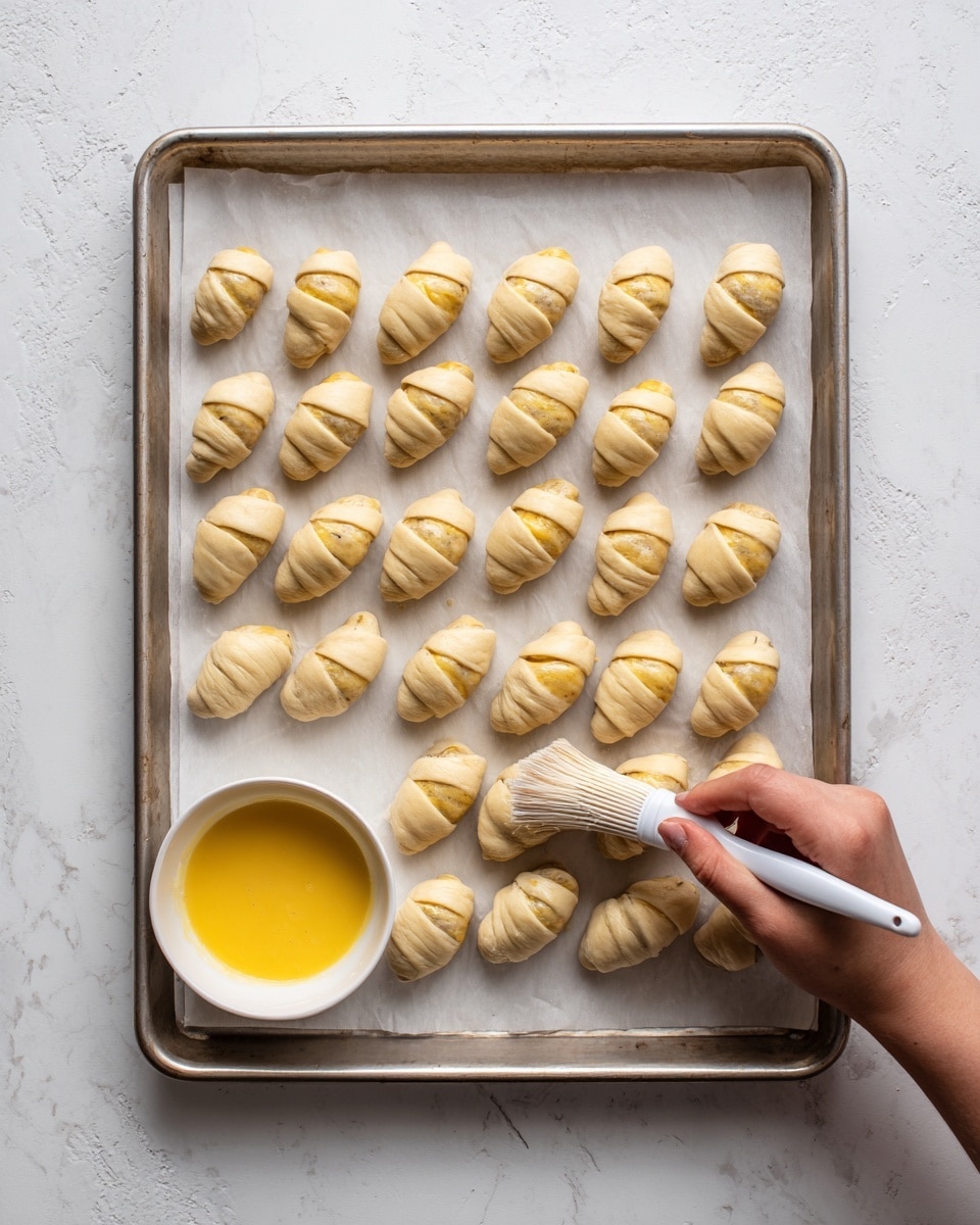 The image shows a silver baking tray lined with white parchment paper, holding 24 small sausages wrapped in light beige dough in neat rows. Each sausage is almost fully covered with the dough, revealing just the ends, and the dough has a smooth, slightly shiny texture. A woman's hand holding a white brush is applying a light yellow egg wash on one of the dough-wrapped sausages near the bottom left corner. To the left of the tray, there is a small white bowl filled with the yellow egg wash. The tray rests on a white marbled textured surface. photo taken with an iphone --ar 4:5 --v 7