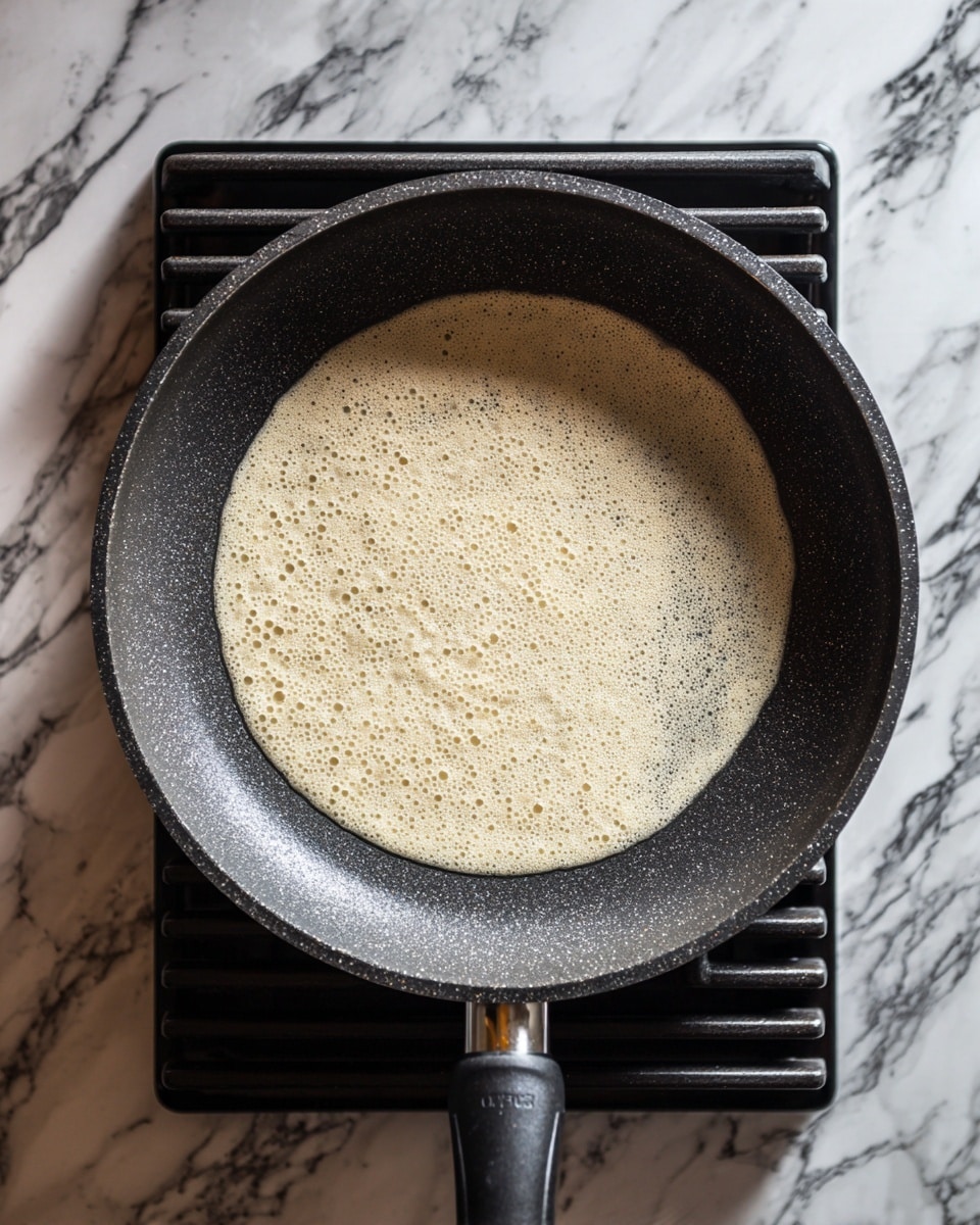 A frying pan with a dark, slightly textured inside surface is shown on a stove with black grates. Inside the pan, there is a thin, light beige pancake batter layer spreading evenly, with some small bubbles forming near the center and edges. The background features a white marbled texture. photo taken with an iphone --ar 4:5 --v 7