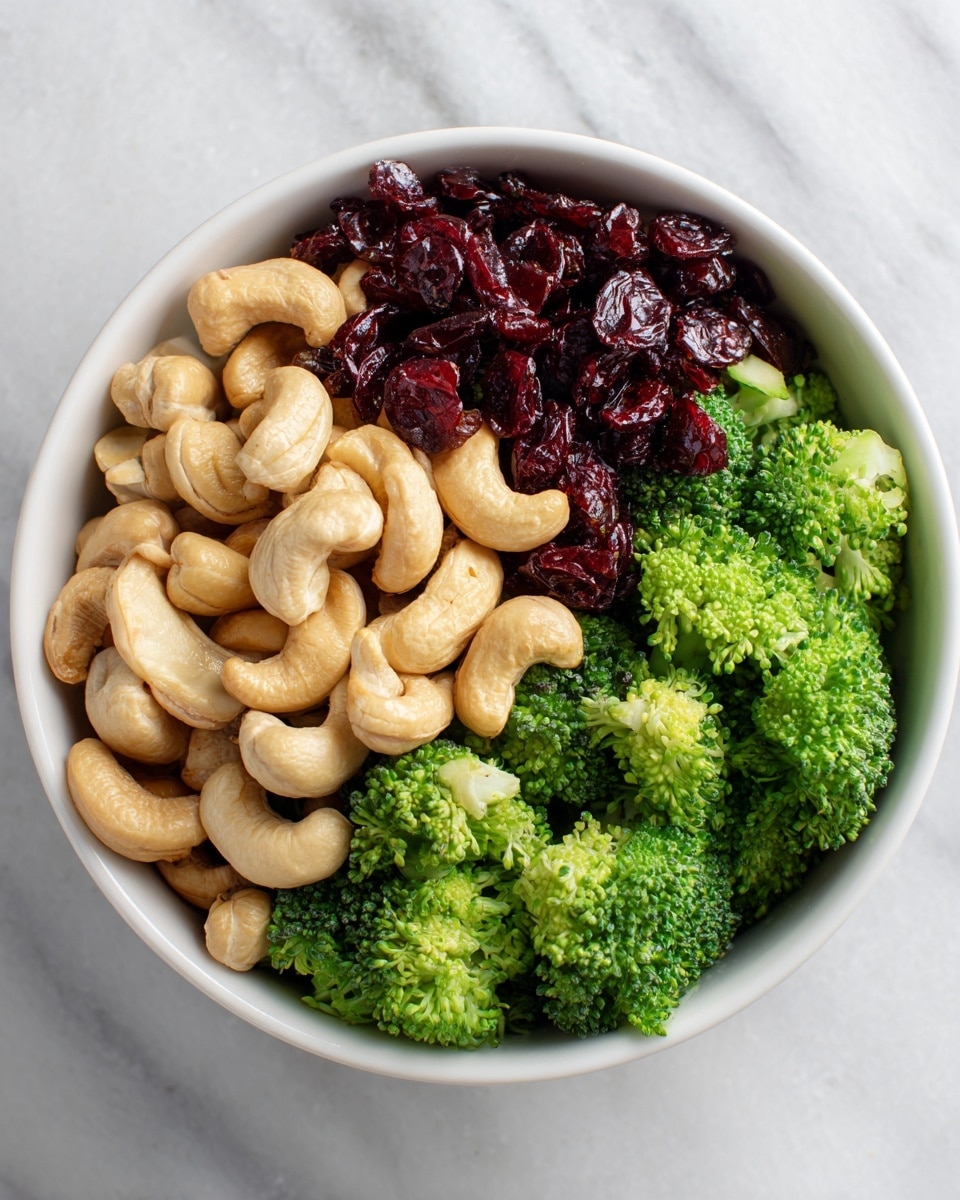 A white bowl filled with fresh, bright green broccoli florets as the base layer, evenly spread out with their textured tops and stalks visible. On top, there is a generous layer of light beige cashew nuts with smooth, curved shapes scattered evenly. The topmost layer consists of dark red dried cranberries, adding a rich contrast in color and a slightly wrinkled texture, scattered across the nuts and broccoli. The bowl rests on a white marbled surface, showing subtle gray veining. Photo taken with an iphone --ar 4:5 --v 7