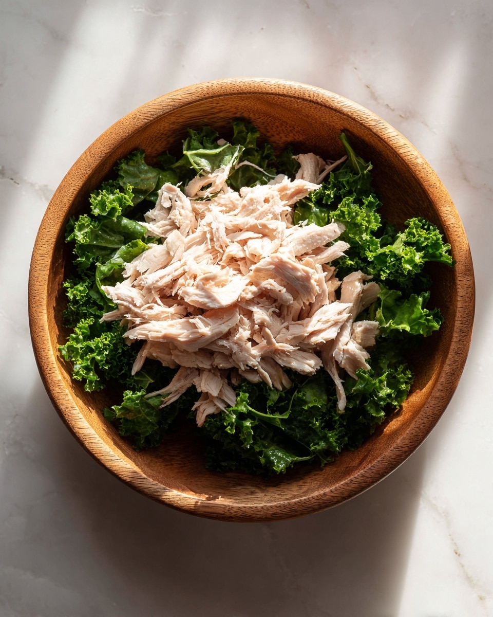 A wooden bowl filled with a base layer of bright green kale leaves, fresh and leafy with a slightly rough texture. On top of the kale, there is a generous pile of shredded pale pink chicken with a soft, fibrous texture, loosely placed in the center. The bowl sits on a white marbled surface with sunlight shining on the food, creating strong shadows and highlights. photo taken with an iphone --ar 4:5 --v 7
