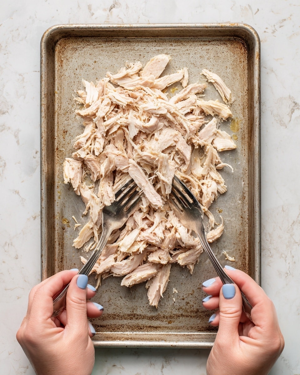 In the image, pieces of cooked white meat are placed in a single layer on a worn metal baking tray that shows some marks and stains from use. The shredded meat varies in shape and size with a slightly golden outer edge and a soft white inside. Two silver forks are lifting and pulling apart the meat, creating texture with visible strands. At the bottom corners, two pale-colored woman's hands with blue nail polish hold the forks. The tray sits on a white marbled surface. photo taken with an iphone --ar 4:5 --v 7