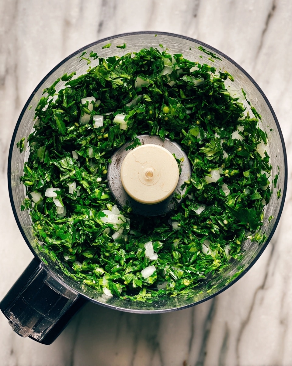 A clear food processor bowl filled with finely chopped bright green herbs and small white pieces, evenly mixed inside. The white blade sits in the center, surrounded by the green and white mixture. The bowl is placed on a white marbled surface, and the black handle of the food processor is visible on the left side. photo taken with an iphone --ar 4:5 --v 7