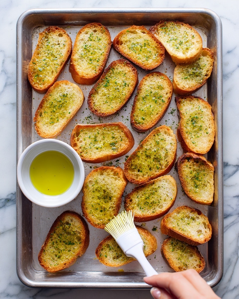 The image shows a metal baking tray with 18 slices of toasted bread arranged all over the tray. Each bread slice is golden brown with a crispy crust and has a light greenish shine from olive oil brushed on top. A woman's hand is holding a small white basting brush applying more oil to one slice near the bottom center of the tray. On the left side of the tray, there is a small white bowl filled with olive oil. The background is a white marbled surface. photo taken with an iphone --ar 4:5 --v 7