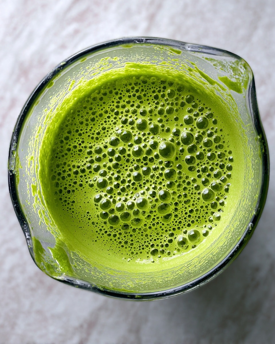A top view of a glass blender filled with bright green smoothie mixture that has many small bubbles on its surface. The blender shows clear sides with some green residue clinging to the inside walls. The background has a white marbled texture. photo taken with an iphone --ar 4:5 --v 7