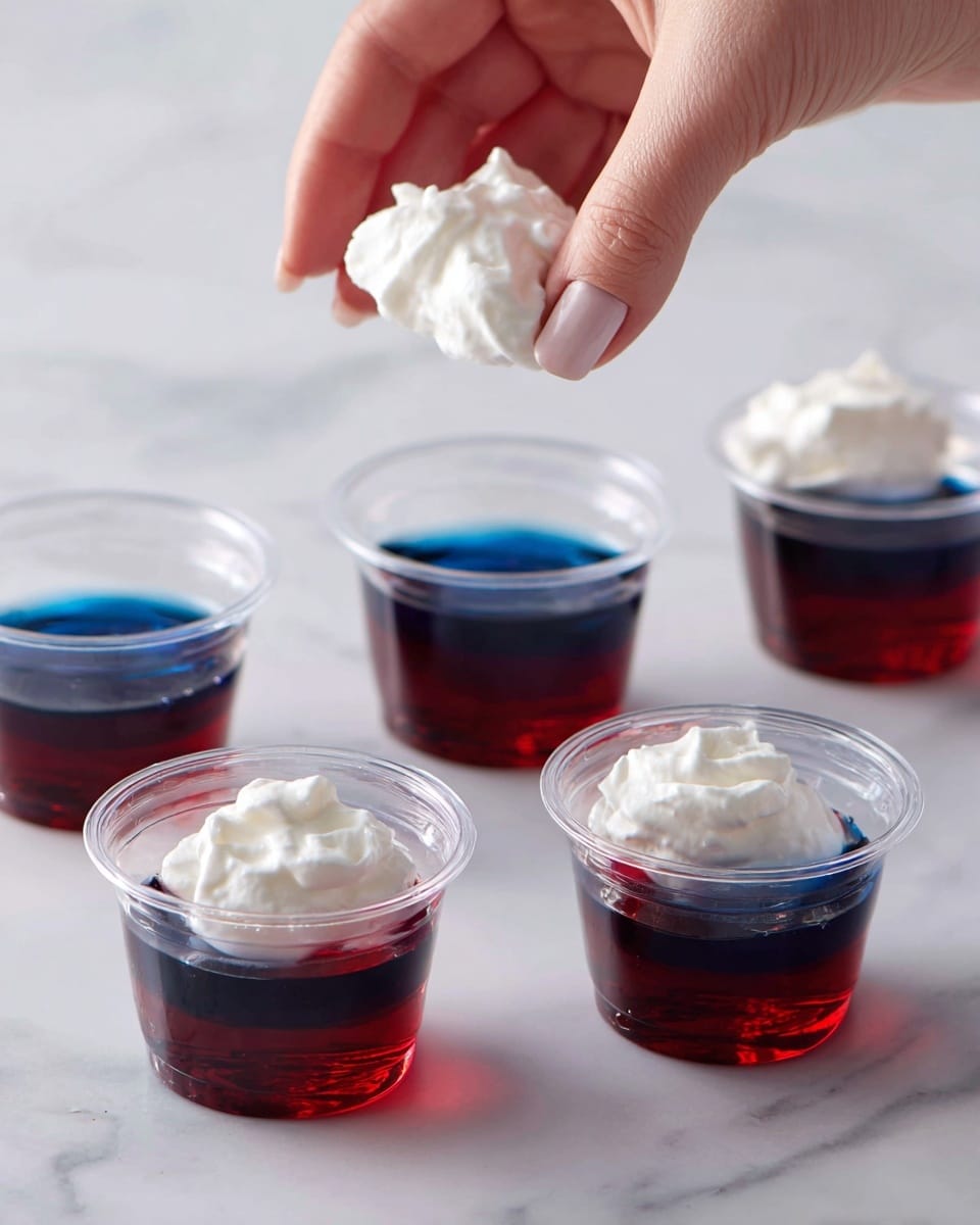 The image shows small clear plastic cups filled with two layers: the bottom layer is a smooth, shiny deep blue jelly, while the top layer is a dark red jelly with a glossy finish. In the second part of the image, a woman's hand is adding a dollop of soft white whipped cream on top of the red jelly layer. The cups are placed on a white marbled surface, creating a clean and bright background. Photo taken with an iphone --ar 4:5 --v 7