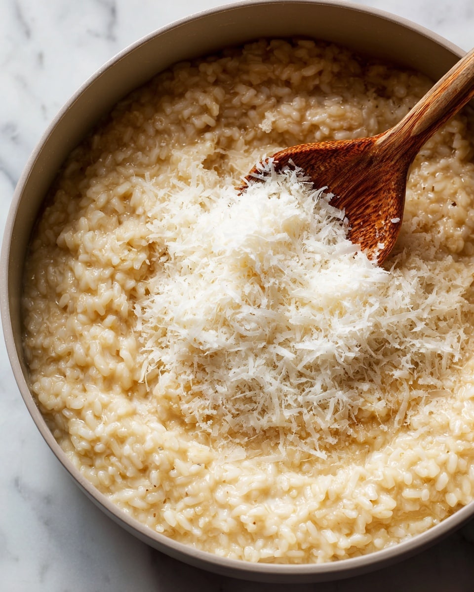 A close-up of a pot filled with creamy risotto made with soft, light beige rice grains. On top, a pile of grated white cheese is being stirred in with a wooden spoon that has a slightly worn texture. The risotto looks rich and smooth, with the rice grains soft and slightly shiny, filling the pot evenly. The background shows a white marbled texture. photo taken with an iphone --ar 4:5 --v 7