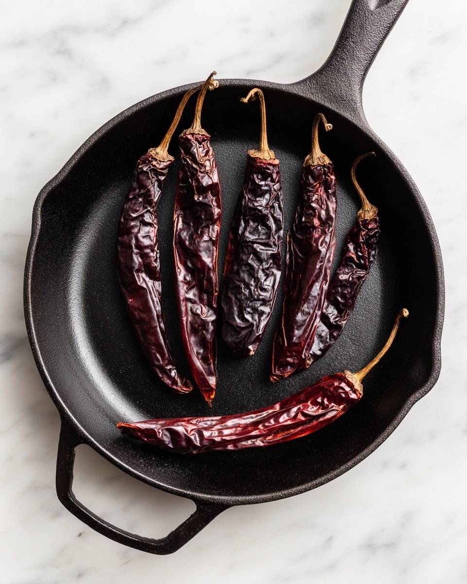 The image shows a black cast iron pan placed on a white marbled surface, containing six dried dark reddish-brown chili peppers of different sizes. They are arranged with four long chilies spread across the center and two smaller, curved ones near the bottom. The chilies have a wrinkled texture with some shiny and slightly charred spots, showing a rough, dry surface. The pan's texture is matte with some light reflections, enhancing the dark color of the chilies and the pan. Photo taken with an iphone --ar 4:5 --v 7