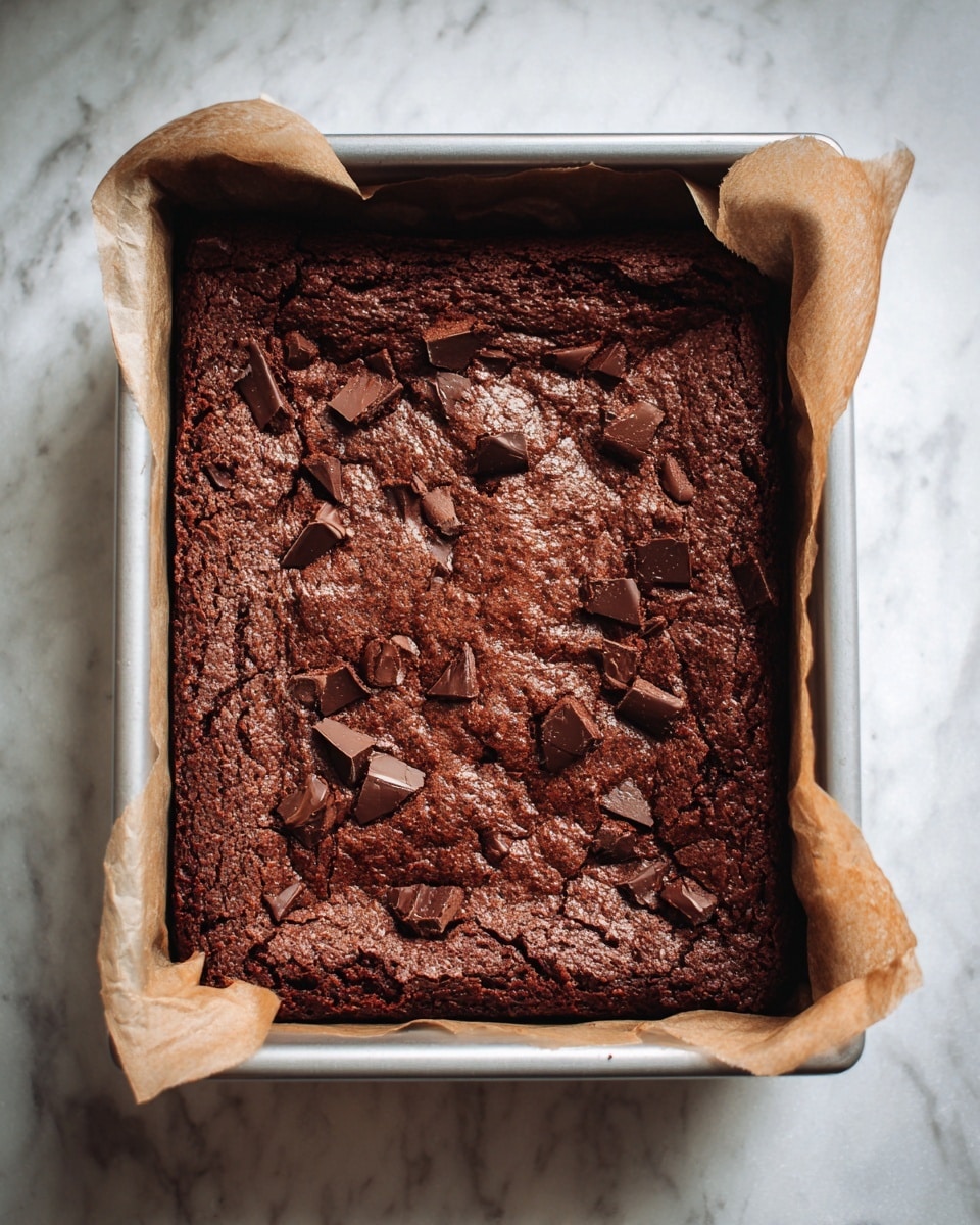 The image shows a freshly baked chocolate brownie in a rectangular metal baking pan lined with parchment paper. The brownie has one thick layer with a rich dark brown color, featuring a slightly cracked and textured surface scattered with small chunks of melted chocolate. The pan rests on a white marbled surface. photo taken with an iphone --ar 4:5 --v 7