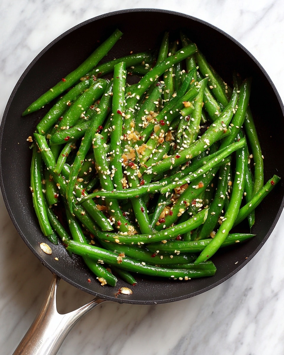 A black pan filled with one layer of bright green cooked green beans, slightly shiny from oil or butter, scattered with small white sesame seeds and bits of red pepper flakes, placed on a white marbled surface. The pan has a metal handle extending towards the bottom left corner of the image. Photo taken with an iphone --ar 4:5 --v 7
