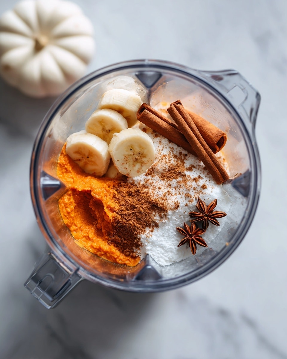 A clear blender jar sits on a white marbled surface with a white pumpkin to the bottom left and two cinnamon sticks and a star anise on the right. Inside the blender are several layers: the bottom layer is bright orange pumpkin puree, above that some white creamy liquid and white powder, and finally some sliced bananas with brown cinnamon powder sprinkled on top. The blade of the blender is visible in the center, partially covered by the ingredients. Photo taken with an iphone --ar 4:5 --v 7