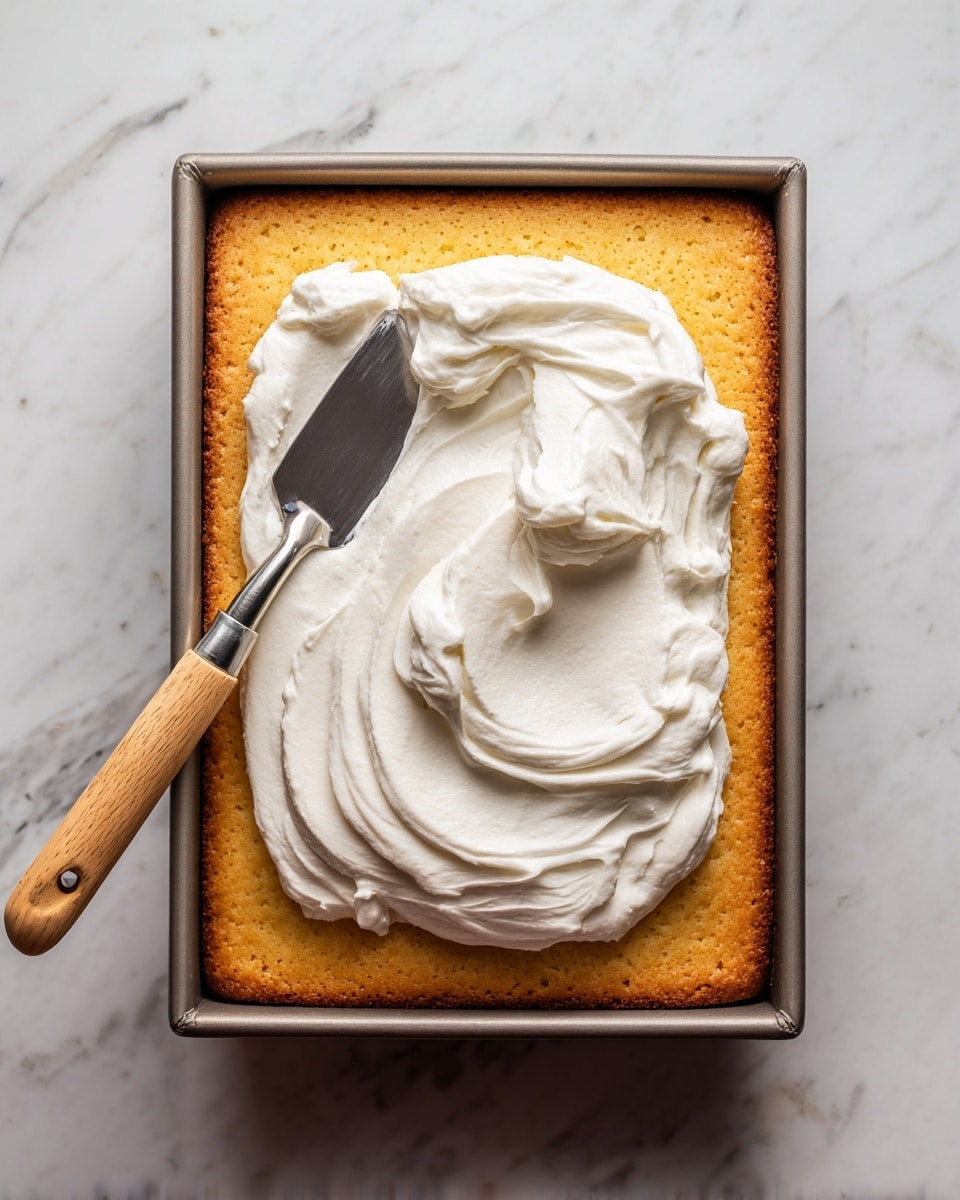 The image shows a single-layer rectangular sponge cake with a golden brown color resting in a baking pan. On top of the cake, thick white whipped cream is being spread using a metal spatula with a wooden handle, creating soft peaks and swirls in the cream. The cake’s surface has small, even holes visible beneath the cream, indicating a light texture. The whole scene is set against a white marbled surface. photo taken with an iphone --ar 4:5 --v 7