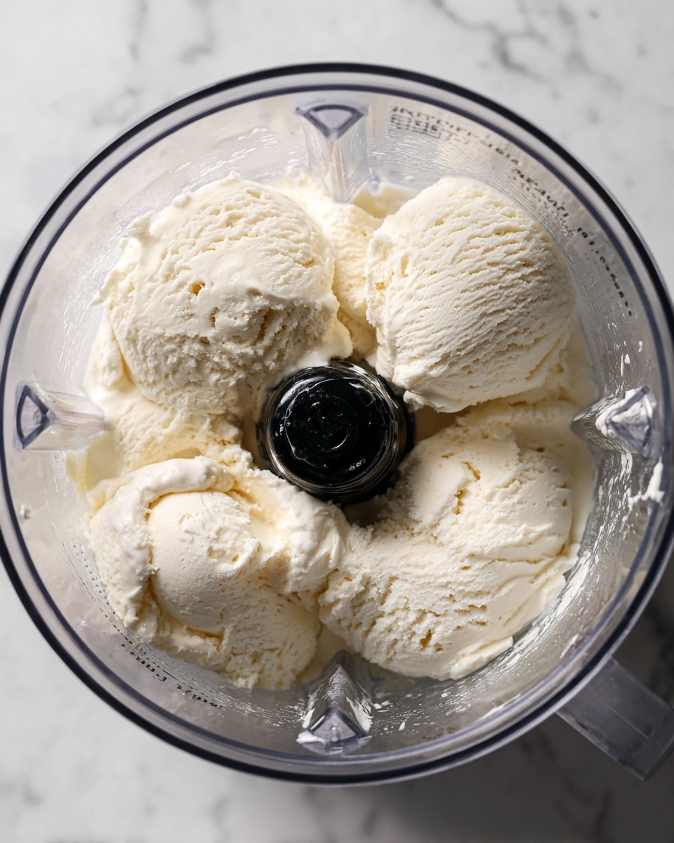 Inside a clear blender container, several large scoops of creamy, white vanilla ice cream fill the space around a black central blade, showing a soft and smooth texture with some small air pockets. The ice cream's surface is slightly shiny and uneven, highlighting its cold and dense quality. The clear container has measurement markings on the side, and the whole blender rests on a white marbled surface. photo taken with an iphone --ar 4:5 --v 7