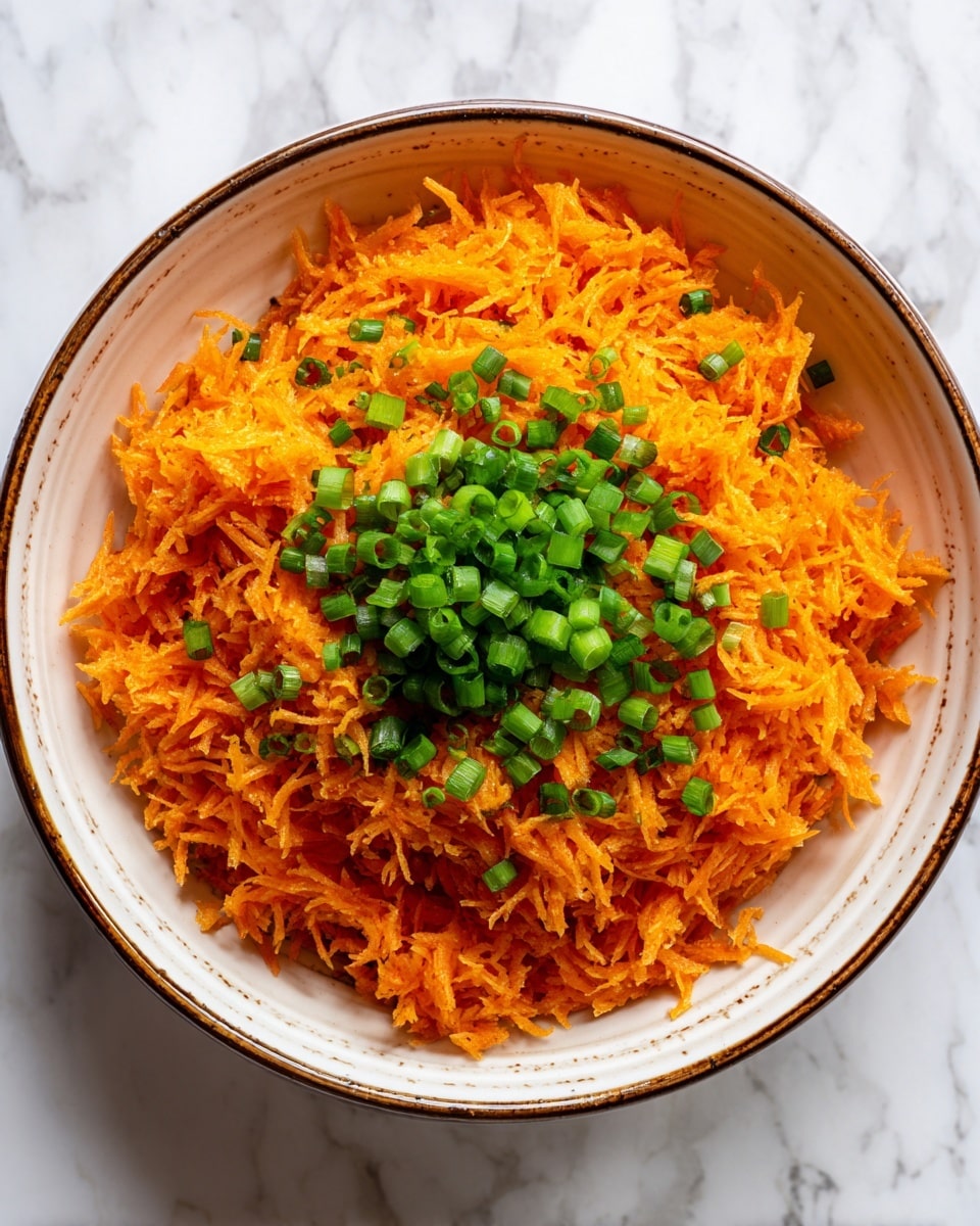 A white bowl with a brown rim filled with two layers: the bottom and main layer is bright orange shredded carrots, finely textured and spread evenly, while a smaller pile of chopped green onions sits neatly on top in the center, adding a pop of deep green color. The bowl is set on a white marbled texture surface. Photo taken with an iphone --ar 4:5 --v 7