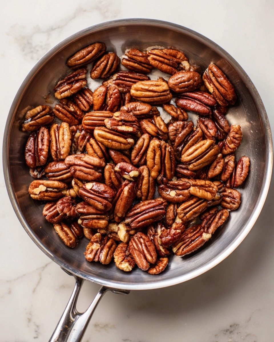 A shiny stainless steel pan filled with many shiny brown pecans that look roasted, showing different shapes and sizes with a smooth texture. The pan is on a white marbled surface, reflecting light softly, and the pecans are scattered evenly inside the pan. photo taken with an iphone --ar 4:5 --v 7