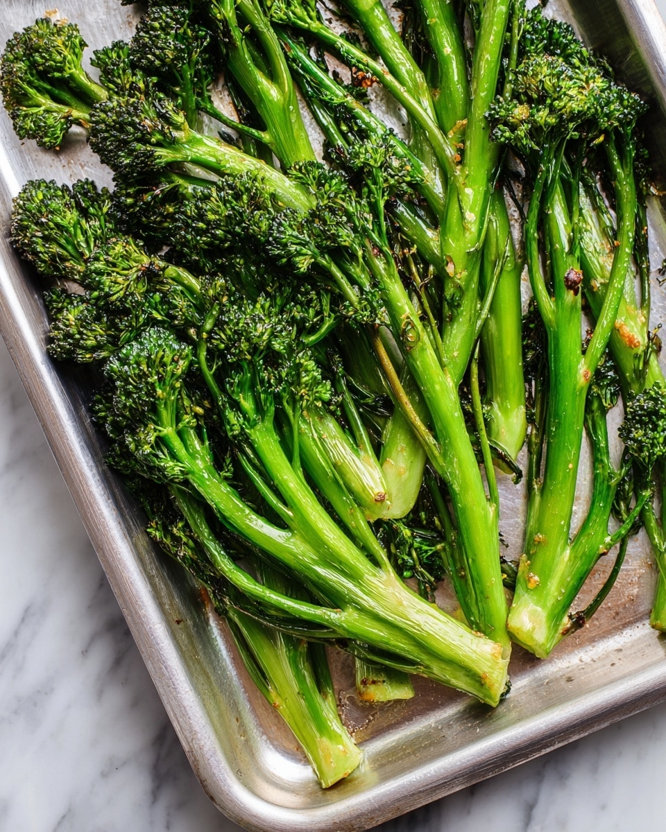 A silver baking tray filled with 11 roasted broccolini pieces arranged in loose rows, each stalk bright green with slightly charred tops and stems, showing a mix of fresh and cooked textures. The broccolini is placed on a white marbled surface, creating a clean and simple background that highlights the vivid green color and light browning on the tender florets and stems. Photo taken with an iphone --ar 4:5 --v 7