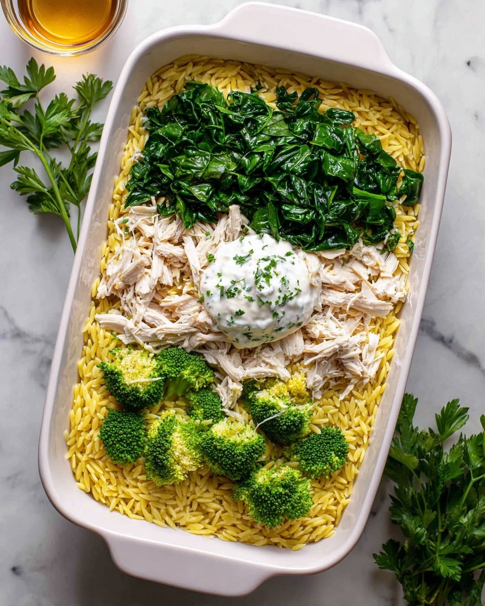 A white rectangular baking dish holds four visible layers of food. The bottom layer is yellow orzo pasta, covering the entire base. On top of the pasta, scattered in different spots, are bright green broccoli florets with a rough, bumpy texture. Over the broccoli, there are pieces of shredded white chicken meat with a soft, fibrous look. The top layer is fresh dark green spinach leaves spread unevenly across the dish. In the center, slightly covered by spinach and chicken, is a round dollop of white creamy sauce with flecks of green herbs. The dish sits on a white marbled surface with a small bunch of parsley and a glass container of light brown liquid in the background. Photo taken with an iphone --ar 4:5 --v 7